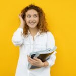 Cheerful young woman standing with books in hand against a vibrant yellow backdrop.